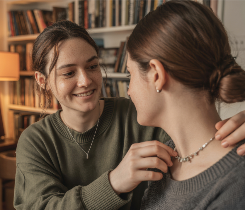 Two people facing each other, one smiling while adjusting the other’s necklace
